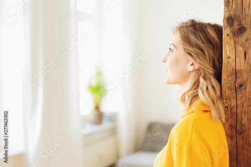 frau lehnt im ihrem wohnzimmer an einem holzbalken und schaut nachdenklich aus dem fenster photo