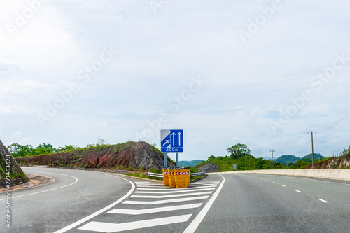 Highway exit with merge/merging lanes ahead street sign/signage and reflective objects on dual carriageway where vehicles drive on left hand side. Scenic commute journey through countryside mountains.