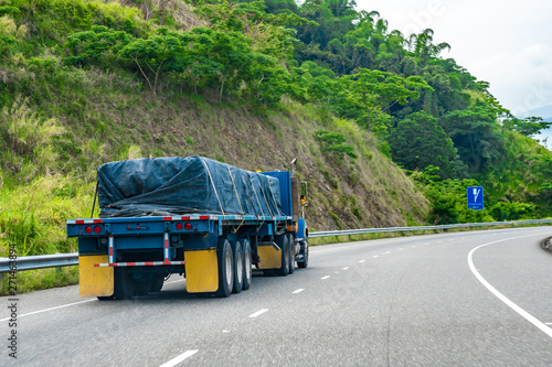 Loaded 22 wheeler cargo semi trailer truck covered and tied/strapped down with rope driving on left hand side of road on dual carriageway. Red and white reflective stickers/ reflectors on rear end.