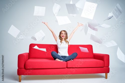 Portrait of pretty young woman throwing stack of papers while sitting on red sofa over white background.