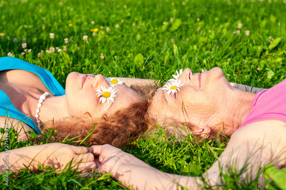 Happy old mother with an adult daughter lie on a glade putting white chamomile flowers to her eyes holding hands. Happy relationship of older mom and daughter. - obrazy, fototapety, plakaty 