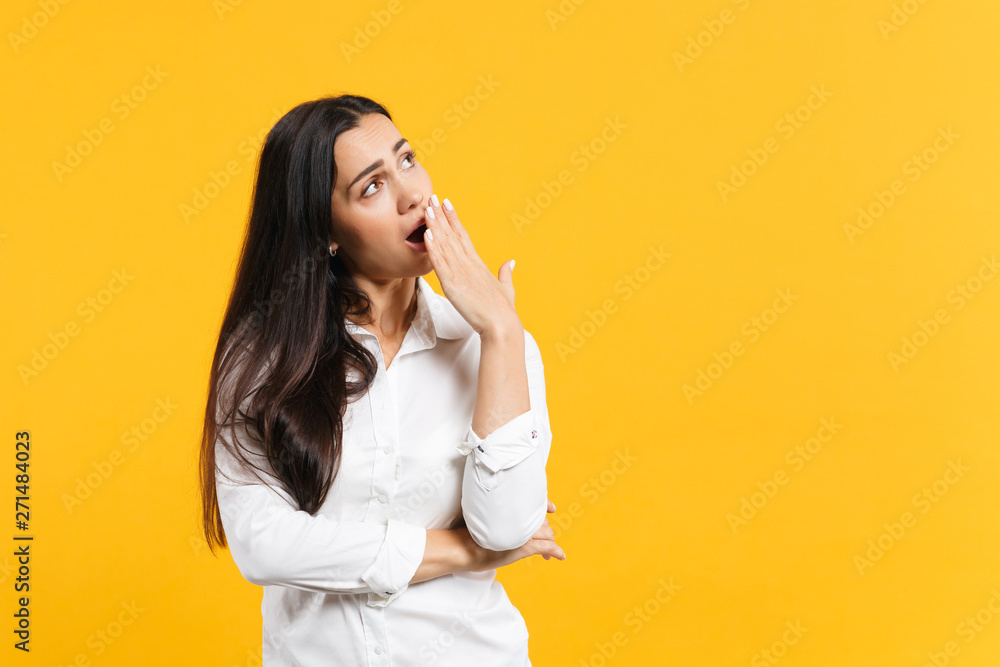Portrait of boring tired young woman in white casual shirt looking aside covering mouth with hand, yawning isolated on yellow orange background in studio. People lifestyle concept. Mock up copy space. Portrait of boring tired young woman in white casual shirt looking aside covering mouth with hand, yawning isolated on yellow orange background in studio. People lifestyle concept. Mock up copy space.