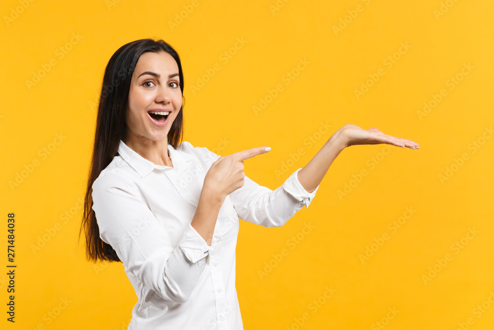 Portrait of excited young woman in white shirt keeping mouth open, pointing index finger, hand aside isolated on yellow orange wall background in studio. People lifestyle concept. Mock up copy space. Portrait of excited young woman in white shirt keeping mouth open, pointing index finger, hand aside isolated on yellow orange wall background in studio. People lifestyle concept. Mock up copy space.