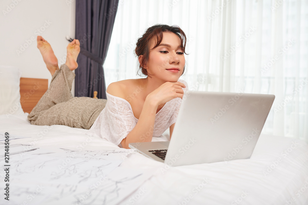Young pretty woman lying on bed and looking at laptop monitor while working with fashion sketches at home Young pretty woman lying on bed and looking at laptop monitor while working with fashion sketches at home