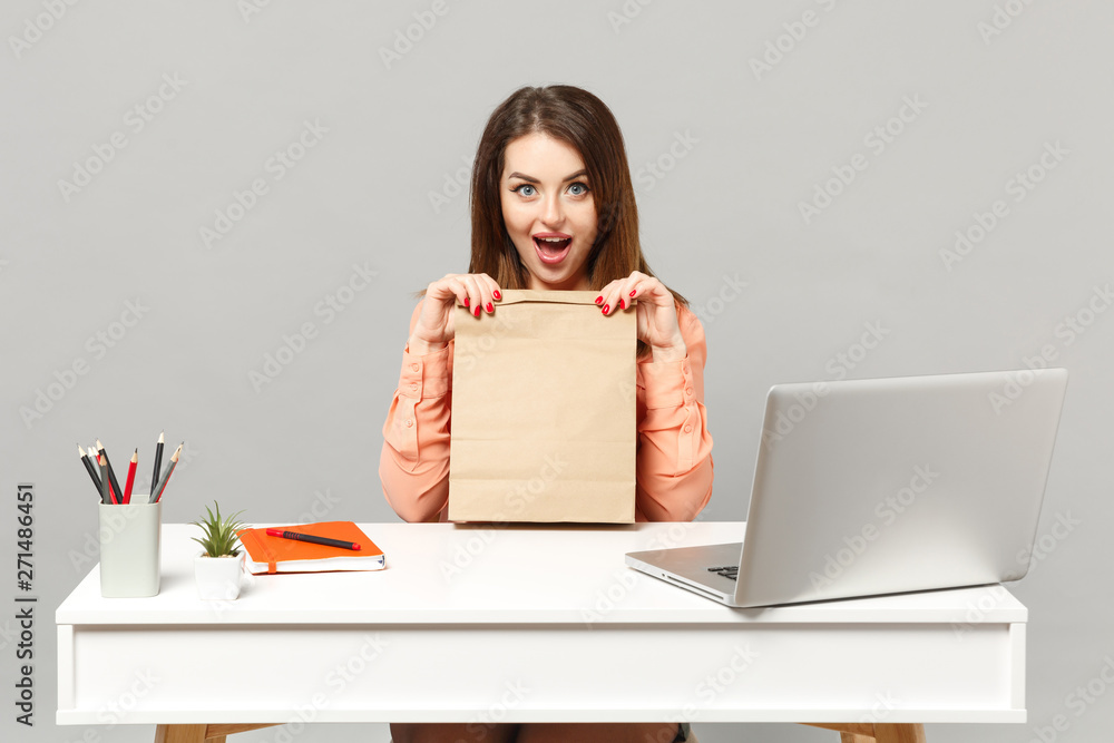 Young excited woman in pastel clothes holding brown clear empty blank craft paper bag for takeaway work at desk with laptop isolated on gray background. Achievement business career lifestyle concept. Young excited woman in pastel clothes holding brown clear empty blank craft paper bag for takeaway work at desk with laptop isolated on gray background. Achievement business career lifestyle concept.