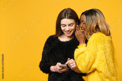 фотографія Two young women friends european and african american in black yellow clothes standing posing isolated on bright orange wall background, studio portrait