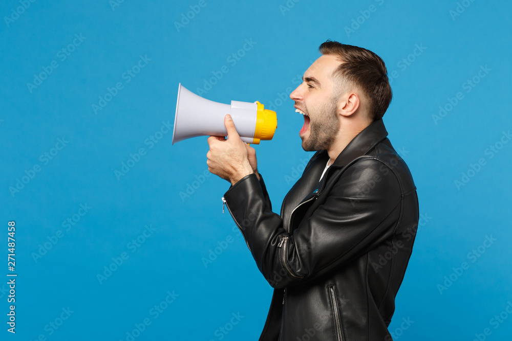 Side view young unshaven man in black jacket white t-shirt scream in megaphone, announces discounts sale isolated on blue wall background studio portrait. People lifestyle concept. Mock up copy space. Side view young unshaven man in black jacket white t-shirt scream in megaphone, announces discounts sale isolated on blue wall background studio portrait. People lifestyle concept. Mock up copy space.