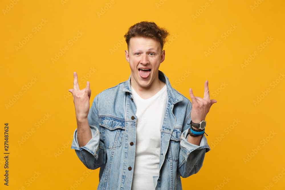 Crazy young man in denim clothes showing tongue, horns up gesture, depicting heavy metal rock sign isolated on yellow orange background. People sincere emotions, lifestyle concept. Mock up copy space. Crazy young man in denim clothes showing tongue, horns up gesture, depicting heavy metal rock sign isolated on yellow orange background. People sincere emotions, lifestyle concept. Mock up copy space.