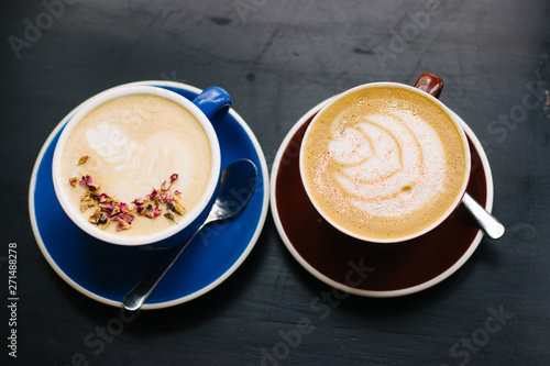 Two cups of cappuccino with latte art on black background. Beautiful foam, blue and brown ceramic cups, stylish toning, place for design and text.