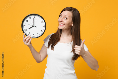 Portrait of pretty young woman in white casual clothes showing thumb up, holding in hand round clock isolated on yellow orange wall background in studio. People lifestyle concept. Mock up copy space.