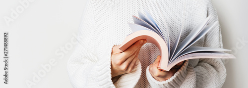 Close view of woman in white woolen sweater holding an open book with pink cover in hands. Long wide banner with free space for your mock up of reading book concept background.