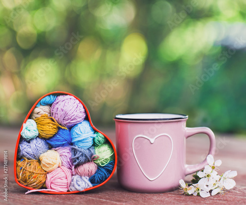 Crochet and knitting. Women's working space. Heart-shaped box with knitted multi-colored skeins of yarn and cup with heart in garden on spring day.