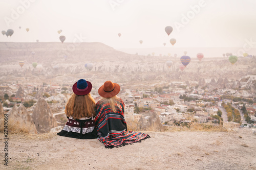 Traveling women wearing authentic boho chic style poncho, sweeter and hats looking on air ballons in sky in Cappadocia valley. Travel and wanderlust concept. Copy space background. photo