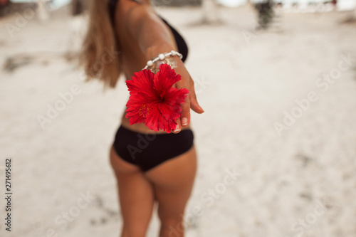 Young tanned woman with perfect buttocks with tattoo on her hand and red flower is walking on the beach. Beauty and fashion concept. © _chupacabra_