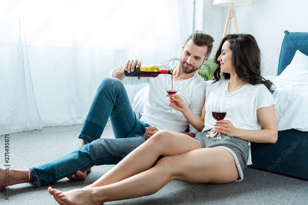Obraz premium boyfriend sitting with girlfriend on floor in bedroom and pouring red wine from bottle in glasses