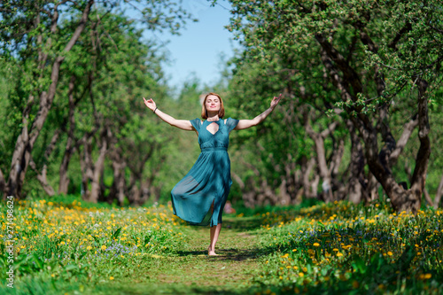 Full-length photo of woman with closed eyes and raised hands in long green dress doing yoga in forest