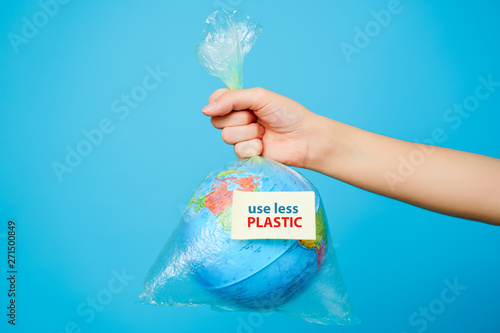Bild auf Leinwand Woman holds in hands plastic bag and planet earth with text sticker- Use LESS PLASTIC at blue background