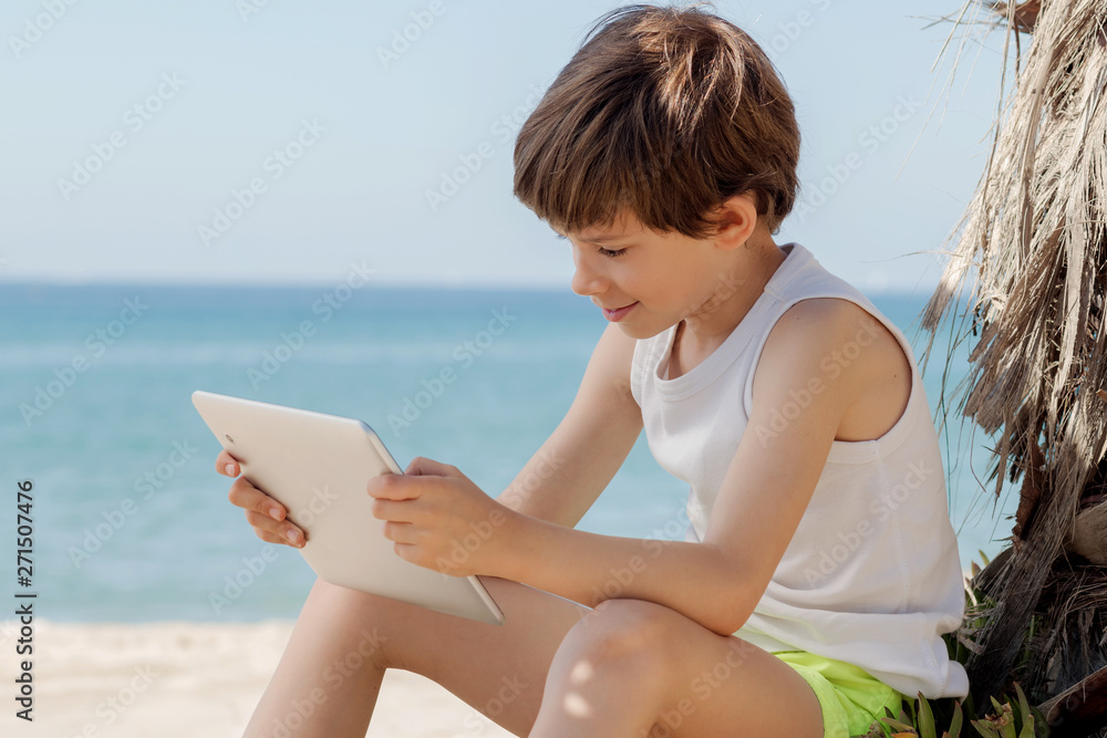 Child looks at tablet in the shade of a palm tree on the beach