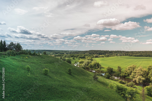 Colorful Bright Sunny Green Field Landscape With Blue Cloudy Sky, River, Trees And Hills