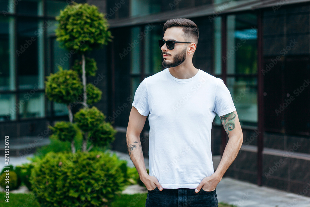 Fototapeta premium A young stylish man with a beard in a white T-shirt and glasses. Street photo