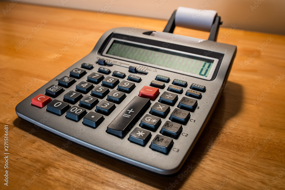 Calculator adding machine on wood desktop. Silver with dark gray buttons, numeric display, and printer paper roll. Calculator adding machine on wood desktop. Silver with dark gray buttons, numeric display, and printer paper roll.