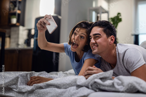 Playful couple making face while taking selfie with smart phone in the bedroom.