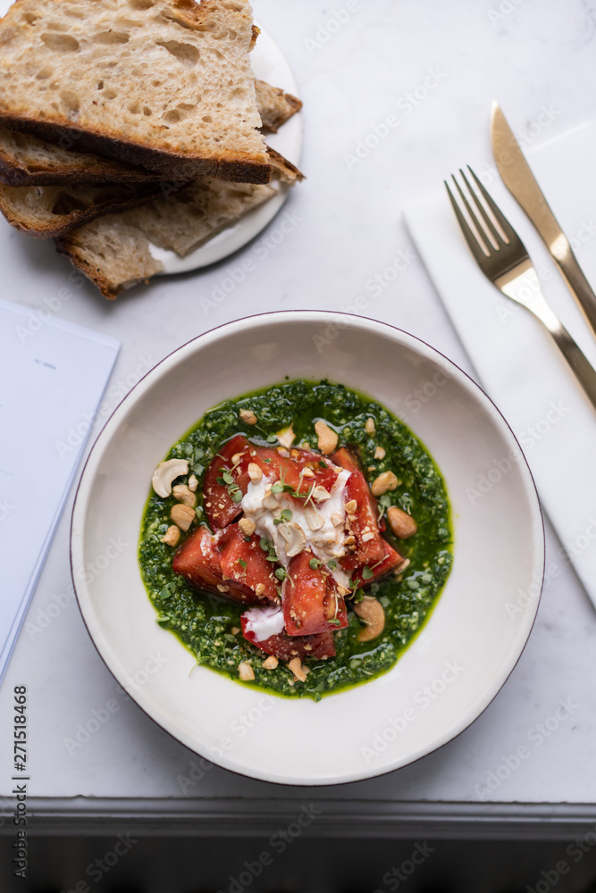 Italian cuisine dish, burrata with fresh red tomatoes in green pesto sauce, olive oil and cashew nuts. All in white bowl with golden cutlery and pieces of black toasted bread. Lunchtime snack in cafe. Italian cuisine dish, burrata with fresh red tomatoes in green pesto sauce, olive oil and cashew nuts. All in white bowl with golden cutlery and pieces of black toasted bread. Lunchtime snack in cafe.