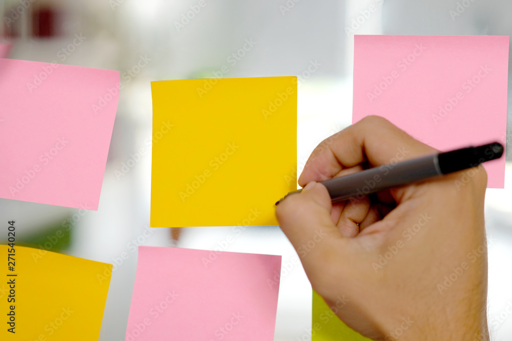 Hand holding pan writing on blank colorful sticky note papers on glass board at office, business stationery supplies Hand holding pan writing on blank colorful sticky note papers on glass board at office, business stationery supplies