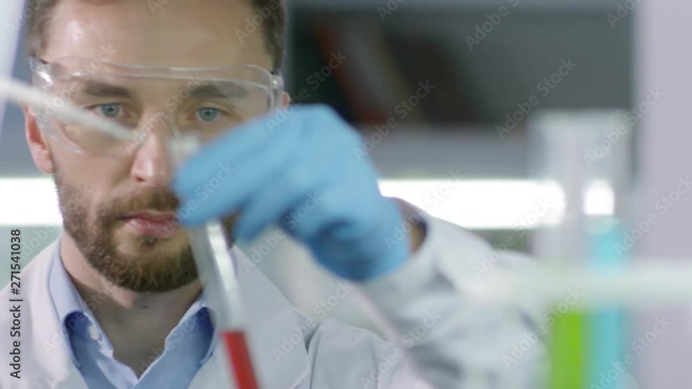 Male scientist in medical clothes and goggles taking red liquid from test tube with pipette Male scientist in medical clothes and goggles taking red liquid from test tube with pipette