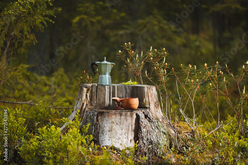 Coffee maker and wooden kuksa stand on a stump in the forest. Coffee maker and cup in focus. The forest background is blurred.