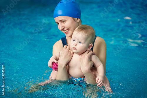Young smiling mother standing to waist in water and holding her little son doing exercise during wellness water treatments for infants in swimming pool. Blue clear water on background. Front view