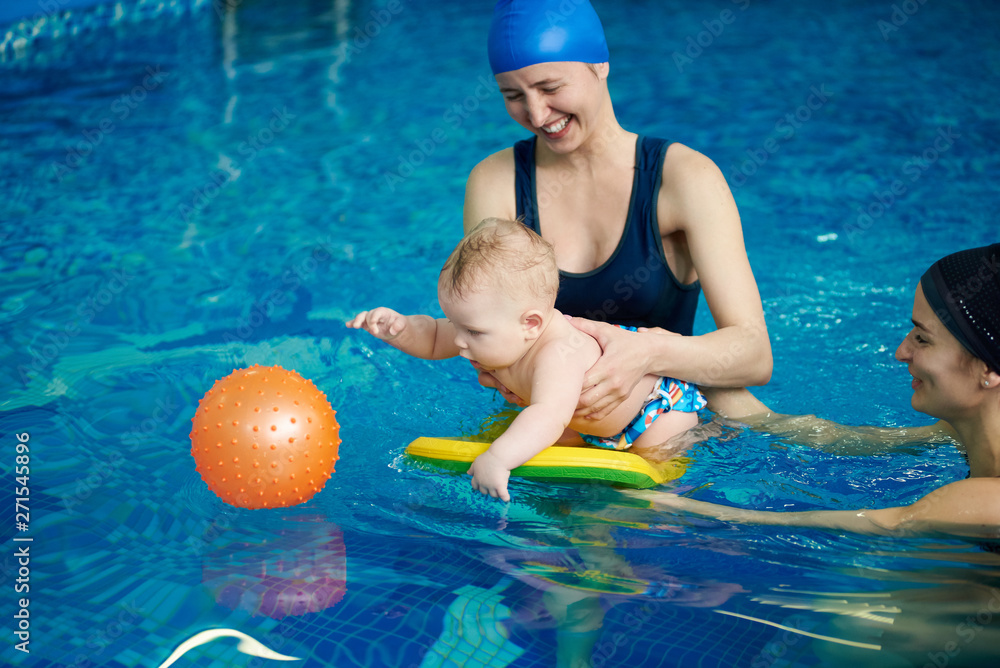 Cute active little child playing in swimming pool. Baby staying on water with board helping and trying getting orange rubber ball in blue water. Family activity and early development concept