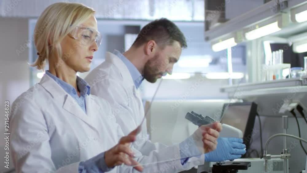 Medium shot of female scientist standing in lab with transparent AR screen in her hands and telling her male laboratory assistant about further working process on microscope Medium shot of female scientist standing in lab with transparent AR screen in her hands and telling her male laboratory assistant about further working process on microscope