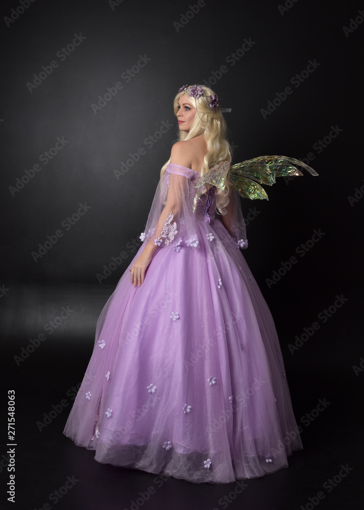 full length portrait of a blonde girl wearing a fantasy fairy inspired costume, long purple ball gown with fairy wings, standing pose with back to the camera on a dark studio background. full length portrait of a blonde girl wearing a fantasy fairy inspired costume, long purple ball gown with fairy wings, standing pose with back to the camera on a dark studio background.