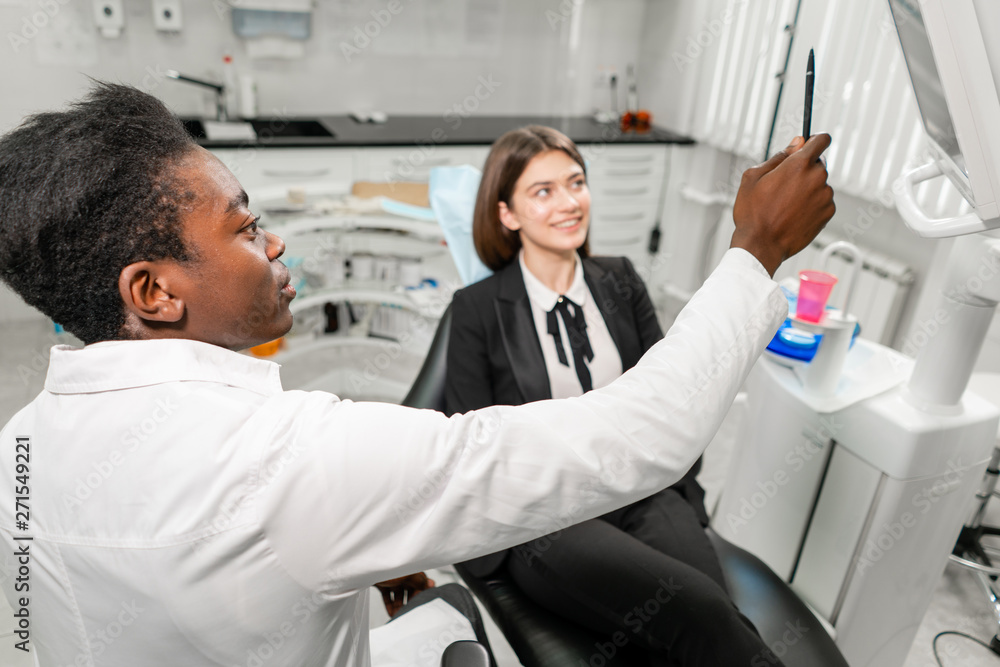 Obraz premium Young African male dentist shows x-rays on the monitor. Woman in the dentist chair at dental clinic. Medicine, health, stomatology concept. dentist treating a patient.