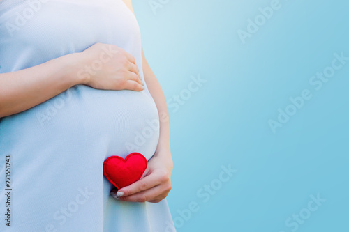 A pregnant woman in a blue dress holds in her hands a red heart on a blue background. Pregnancy, parenthood, motherhood concept. Love for children concept with copy space.