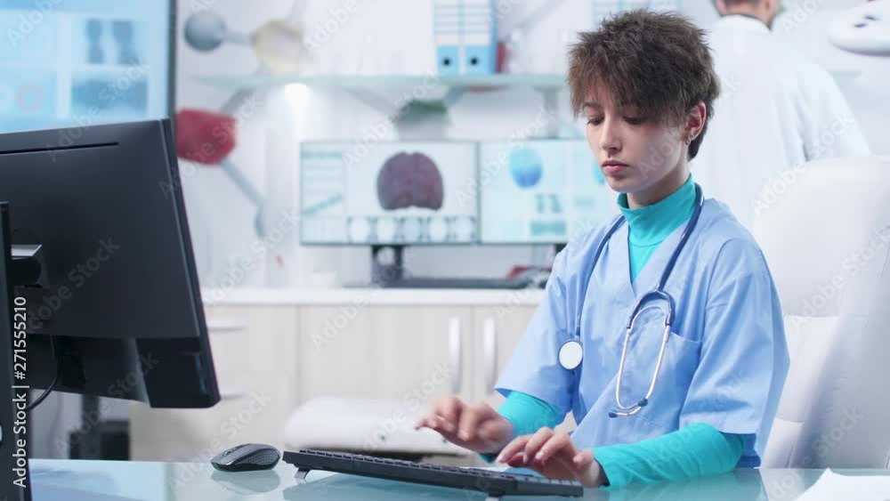 Young nurse typing on computer keyboard in high end tech research center while the doctor is conducting studies on the computers in the background Young nurse typing on computer keyboard in high end tech research center while the doctor is conducting studies on the computers in the background