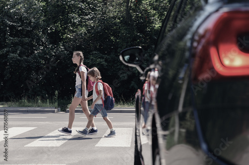 Low angle on car in front of children with backpacks walking through crosswalk to the school