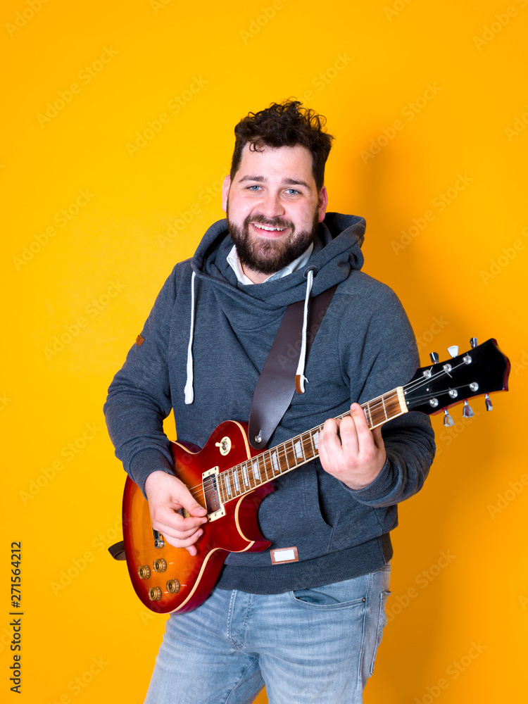 Obraz premium man with black hair and beard, wearing grey hoodie playing the electric guitar in front of a yellow background