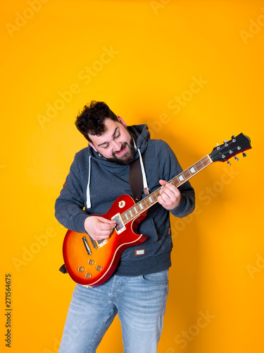 man with black hair and beard, wearing grey hoodie playing the electric guitar in front of a yellow background © epiximages