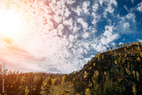 Picturesque landscape of the mountains glowing under sunlight. Fantastic sun rays with blue cloudy sky over the mountain covered with yellow trees. Breathtaking nature image.