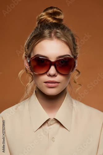 Cropped front view shot of lady with fair hair, wearing beige shirt. The girl with bun and wavy hair locks in oval-shaped sunglasses with brown rim, is looking at camera on light brown background. © RedUmbrella&Donkey