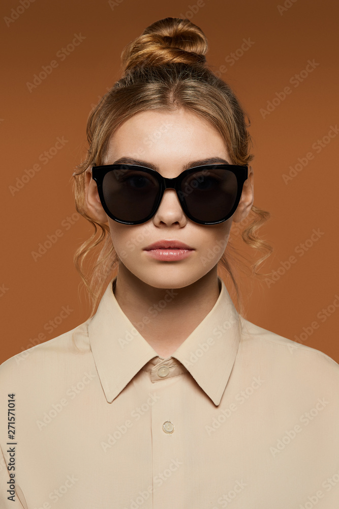Cropped front view shot of blonde lady, wearing shirt. The girl with bun and wavy hair locks in butterfly sunglasses with ink black rim and lenses, is looking at camera against the brown background. Cropped front view shot of blonde lady, wearing shirt. The girl with bun and wavy hair locks in butterfly sunglasses with ink black rim and lenses, is looking at camera against the brown background.