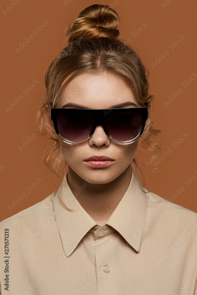 Cropped front view shot of lady, wearing shirt. The girl with bun and wavy hair locks in wrap sunglasses with black gradient rim and dark lenses. The woman is looking at camera over brown background. Cropped front view shot of lady, wearing shirt. The girl with bun and wavy hair locks in wrap sunglasses with black gradient rim and dark lenses. The woman is looking at camera over brown background.