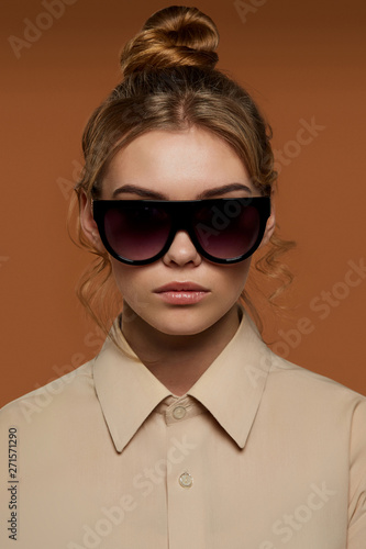Cropped front view shot of lady  wearing shirt. The girl with bun and wavy hair locks in wrap sunglasses with black rim and gradient lenses. The woman is looking at camera against brown background.