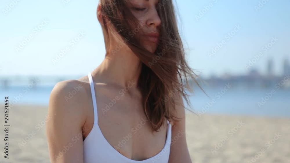 Portrait of a beautiful relaxed young woman sitting in lotus pose at the beach with sea and city background. womans hands in namaste mudra pose. Slim model practice morning meditation on sand city Portrait of a beautiful relaxed young woman sitting in lotus pose at the beach with sea and city background. womans hands in namaste mudra pose. Slim model practice morning meditation on sand city