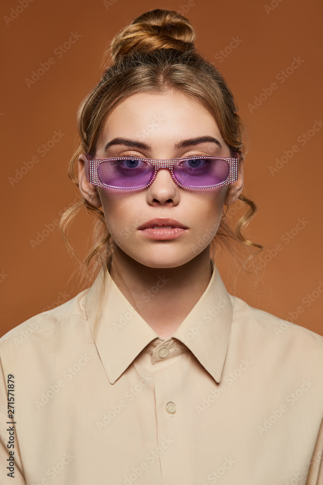 Cropped front view shot of blonde lady, wearing shirt. The girl with bun and wavy hair locks in light purple rectangle sunglasses toreutics on rim, is looking at camera against the sandy background.