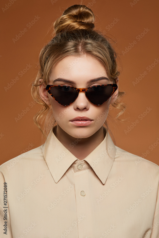 Cropped shot of blonde lady, wearing shirt. The girl with bun and wavy hair locks in cat eye-shaped sunglasses with leopard pattern rim and black lenses, is looking at the camera on brown background.