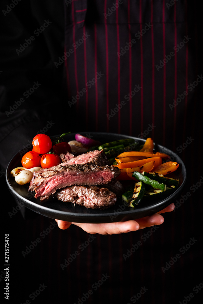 Grilled and sliced beef steak with grilled vegetables served on black plate on black background presentation in chef hands Grilled and sliced beef steak with grilled vegetables served on black plate on black background presentation in chef hands