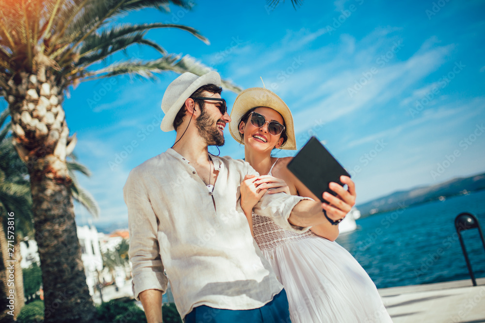 Happy young couple walking by the harbor using digital tablet of a touristic sea resort with sailboats on background